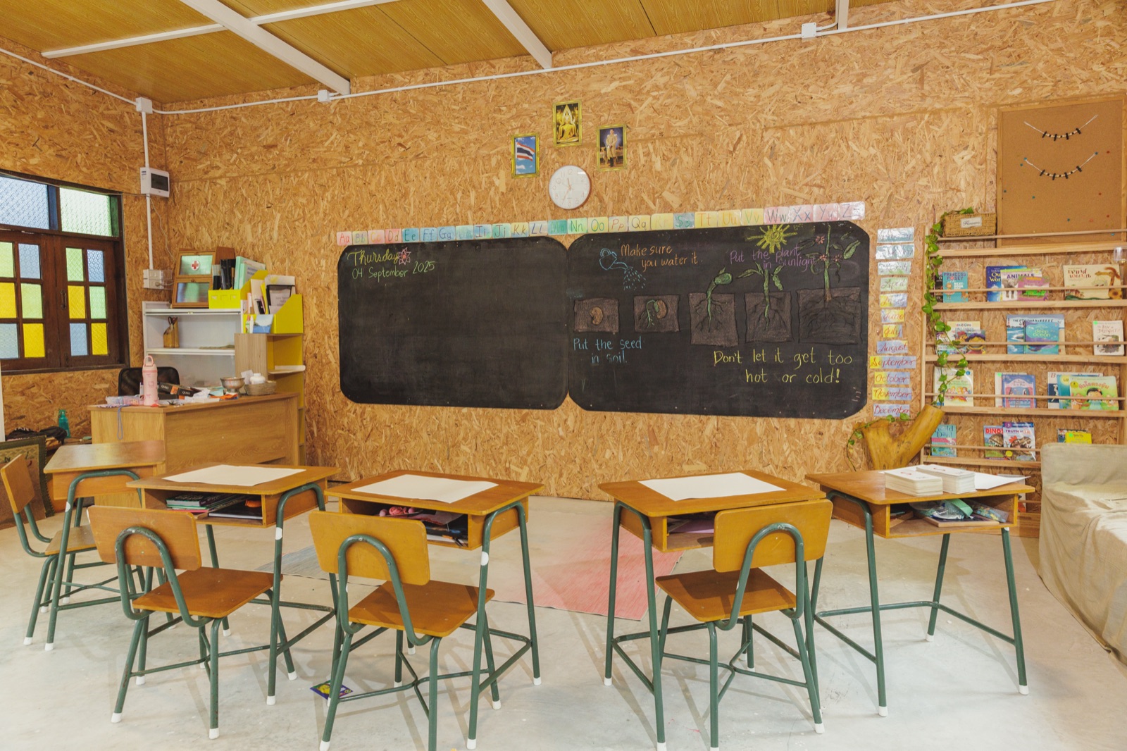 A Bamboo Valley primary classroom with desks facing a chalkboard showing a hand-drawn lesson on how to grow a plant — 'Put the seed in soil. Make sure you water it. Put the plant in sunlight. Don't let it get too hot or cold!'