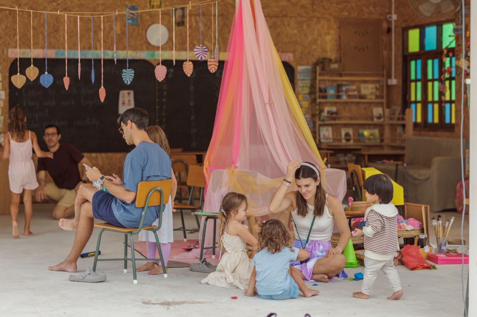 Parents and children inside a Bamboo Valley classroom — children playing together with toys and a soft canopy, the chalkboard behind reading 'Listen to each other, Play and work together, Respect'