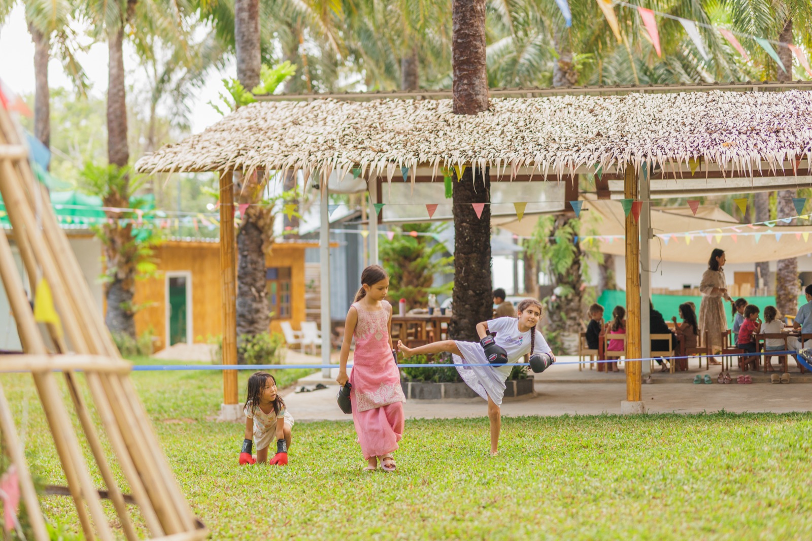 Children with boxing gloves practicing Muay Thai outdoors on the lawn at Bamboo Valley, with the thatched pavilion and other children at tables in the background