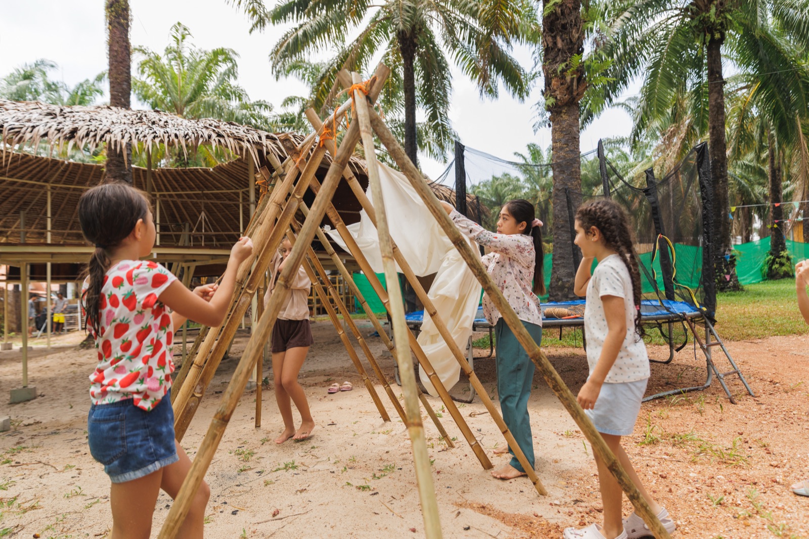 A group of children working together to build a bamboo A-frame fort under the palm trees at Bamboo Valley