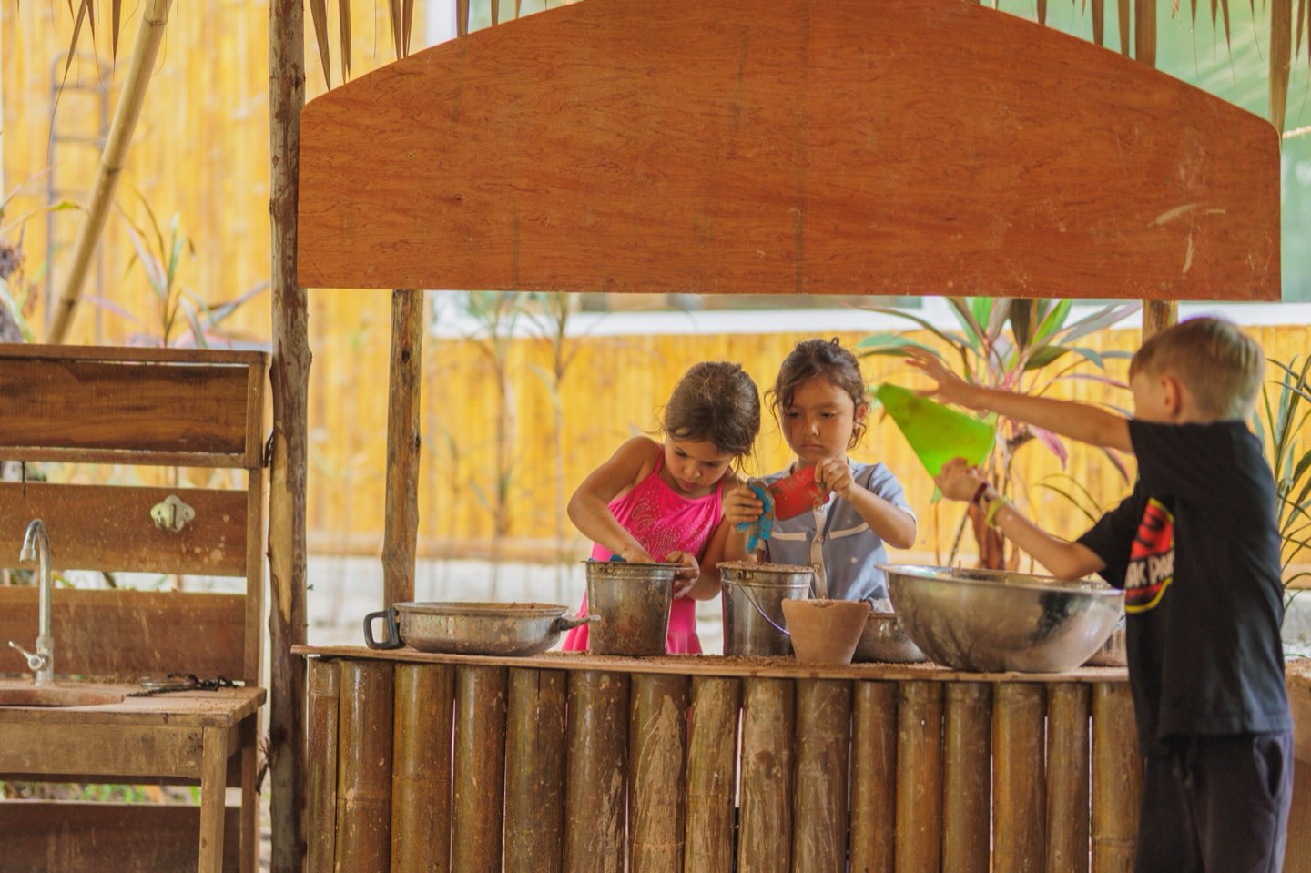 Three young children working side by side at the outdoor mud kitchen at Bamboo Valley, sharing pots and bowls