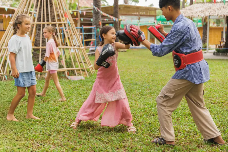 A girl practising Muay Thai punches with a coach on the lawn while friends watch