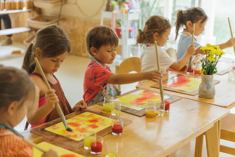 Children painting with watercolours at a wooden table with a vase of yellow flowers