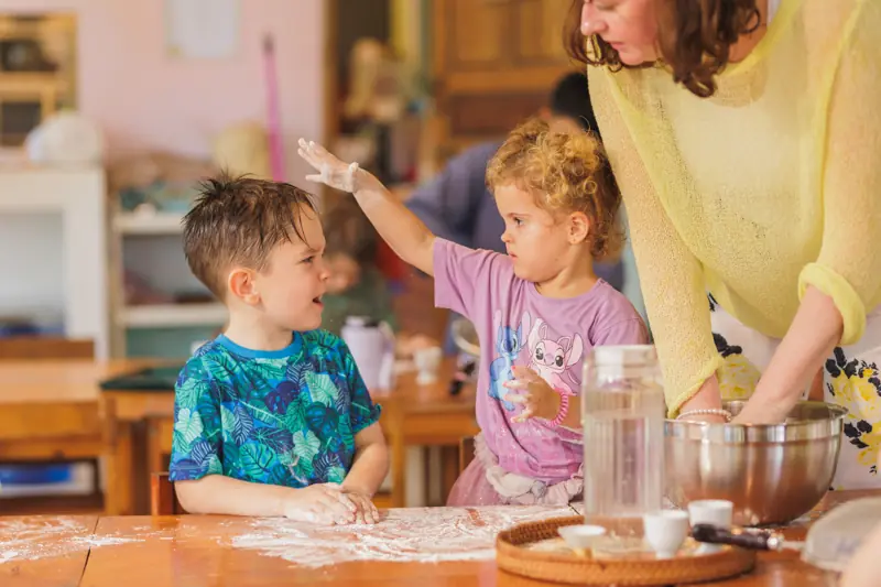 A girl sprinkling flour on a boy at the baking table, both laughing