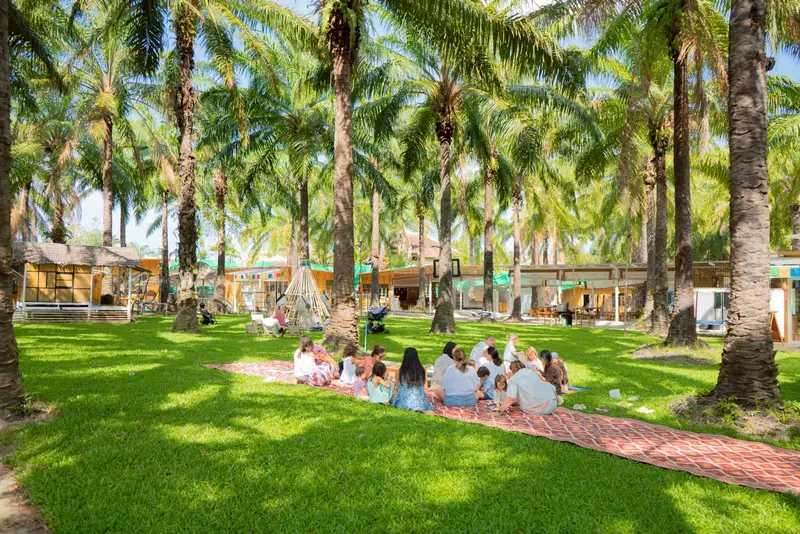 Families and children sitting on a long mat on the lawn in the shade of the palms