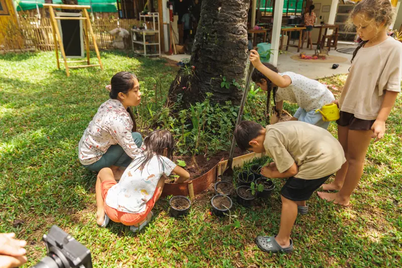 A teacher with children planting a raised bed beside a palm
