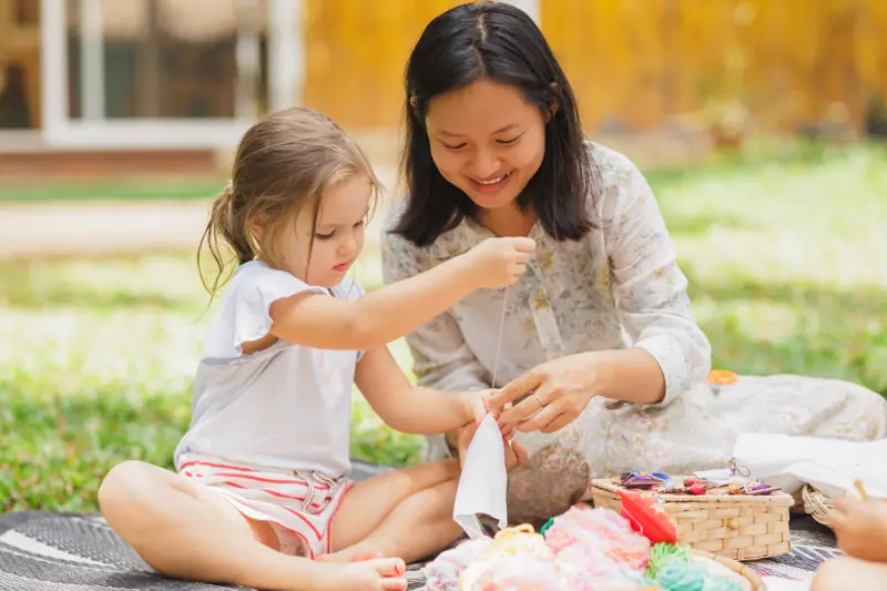A teacher and a girl threading a craft together on a blanket outdoors
